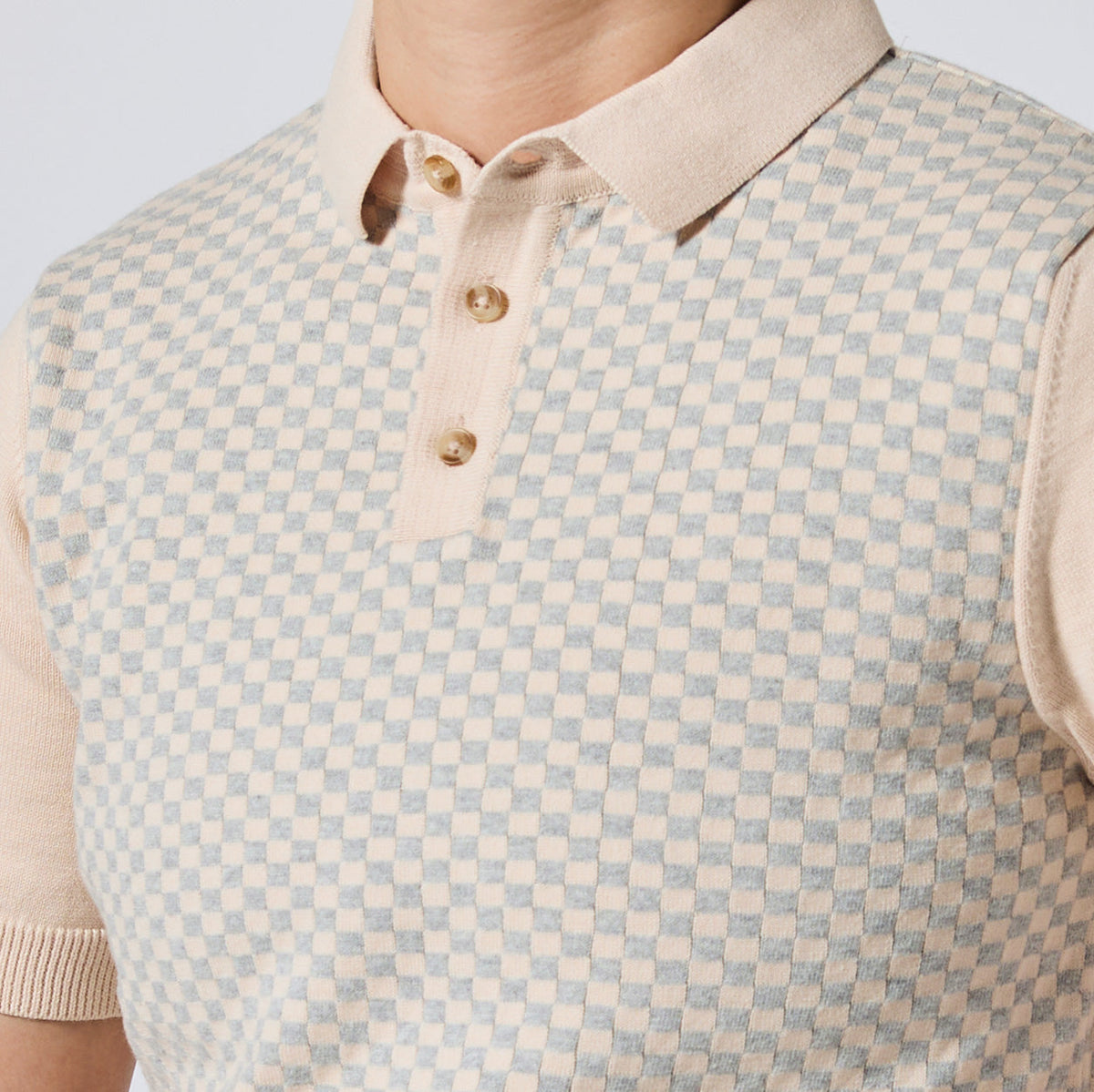 Close-up of a person wearing a short-sleeve knit polo shirt with a beige and light blue checkerboard pattern, a solid beige collar, and three gold buttons. Color: Saltstone Checker