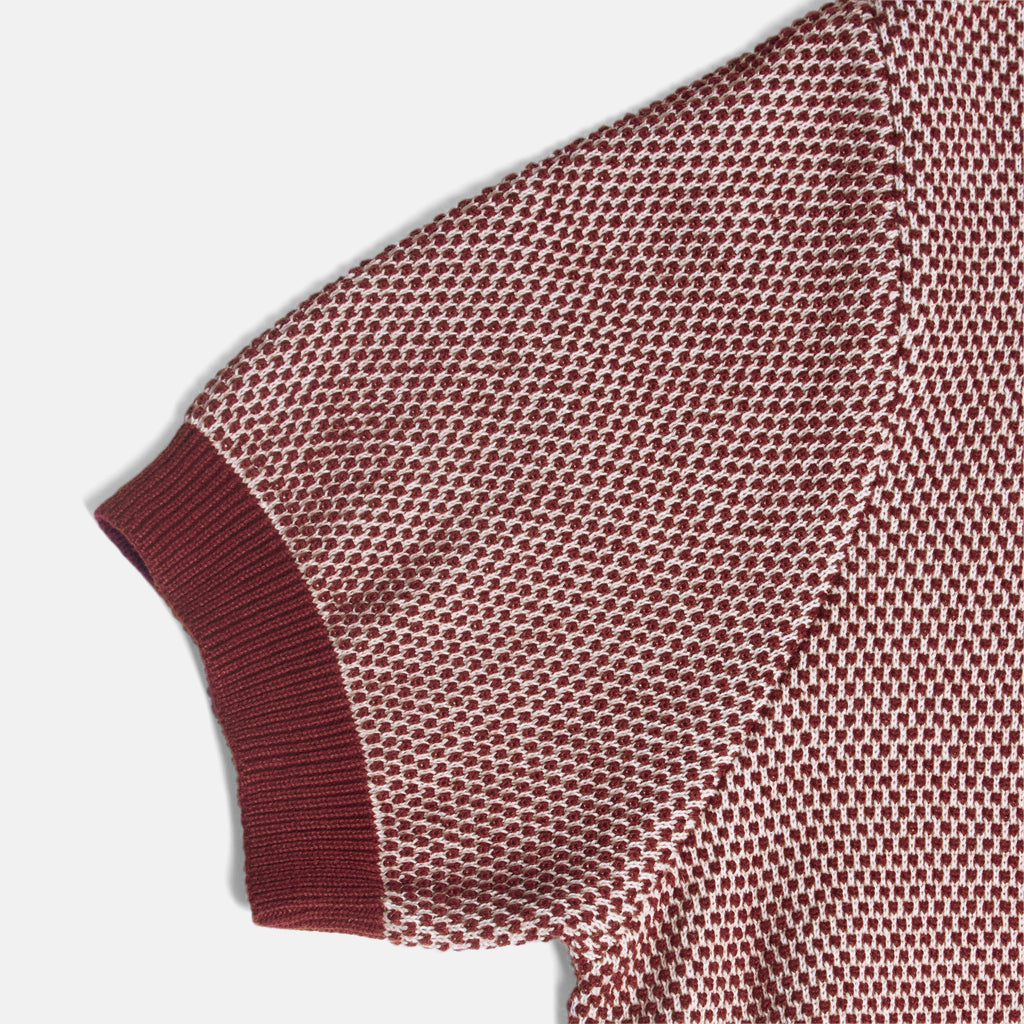 Close-up of a short-sleeve knit top showing the sleeve cuff and textured mesh fabric in a muted burgundy and off-white pattern, on a light gray background. Color: Auburn