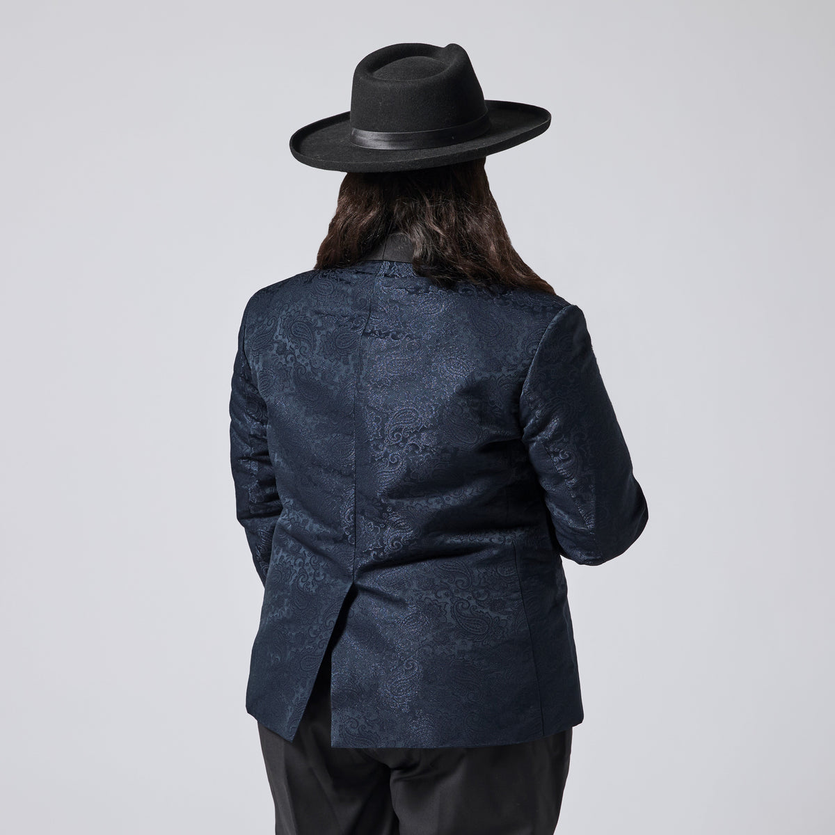 A person shown from the back wearing a dark navy patterned blazer, black pants, and a black wide-brim hat against a light gray studio background. Color: Sapphire Brocade