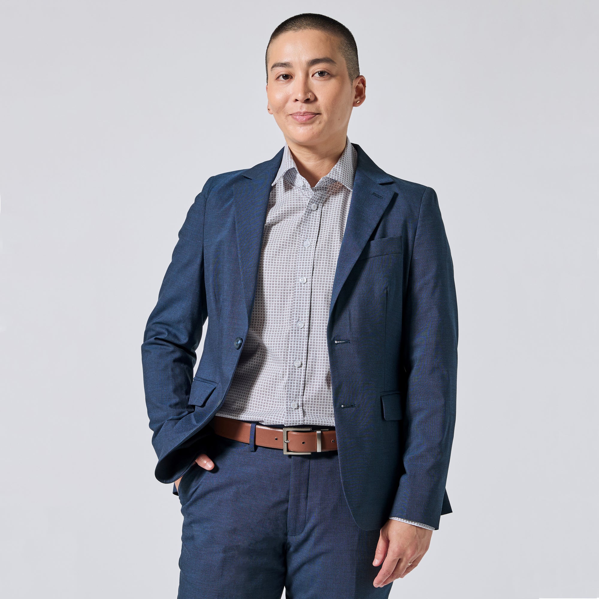 Person wearing an Atlantic blue blazer and matching trousers, paired with a light gray checkered button-down shirt and a brown leather belt, standing against a light gray studio background. Color: Atlantic Blue