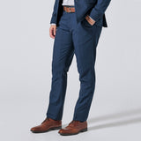 Person wearing Atlantic blue trousers, a matching navy blazer, a white shirt, a brown belt, and brown leather dress shoes, shown from the waist down on a light gray studio background. Color: Atlantic Blue