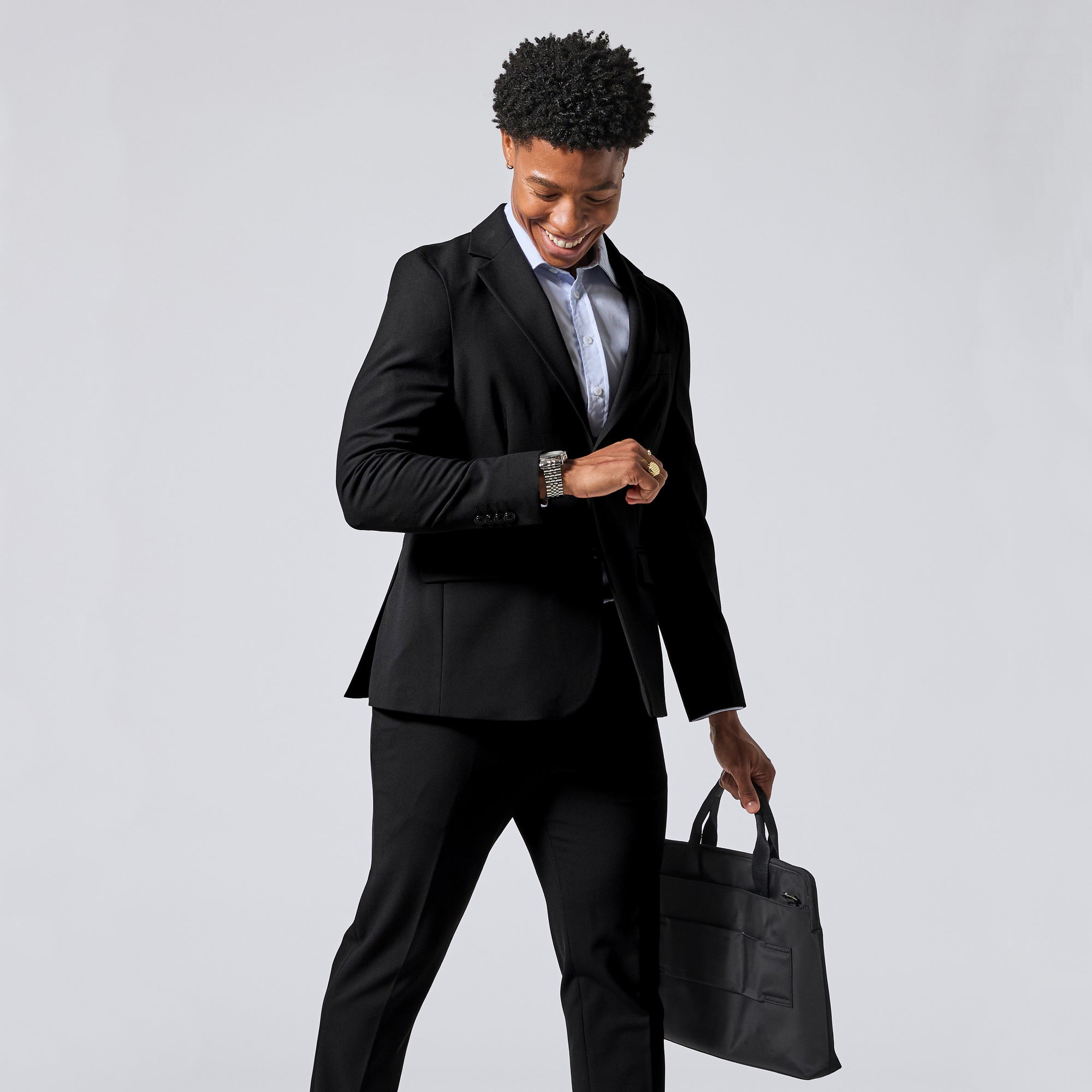 Person wearing a black two-piece suit, light blue dress shirt, and a gold ring, holding a black briefcase against a solid gray background. Color: Obsidian