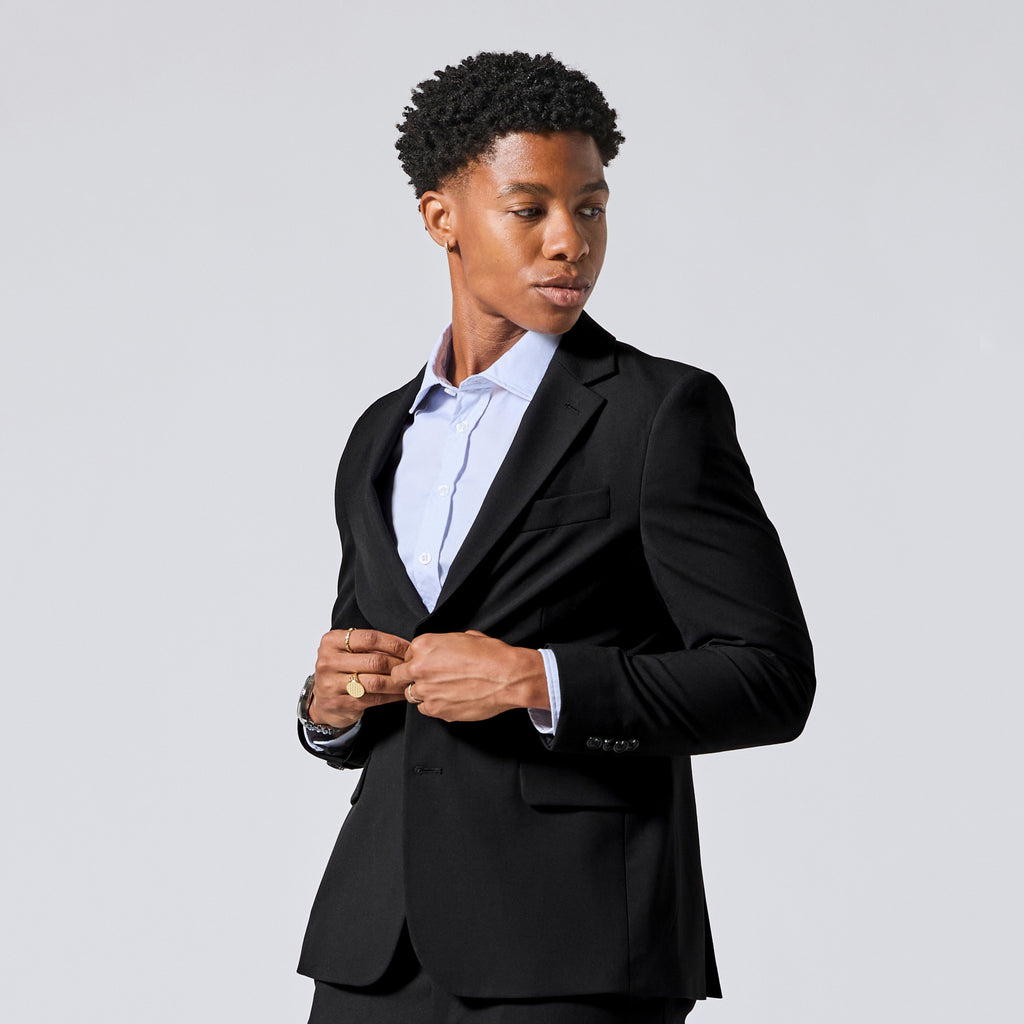 Person wearing a black tailored blazer over a light blue dress shirt, standing against a plain light gray studio background and buttoning his blazer. Color: Obsidian