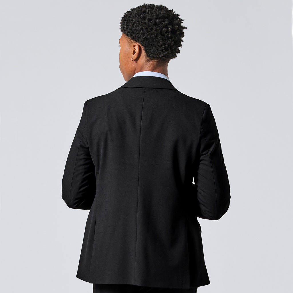 Person viewed from the back wearing a black suit jacket over a white collared shirt, standing against a light gray studio background. Color: Obsidian