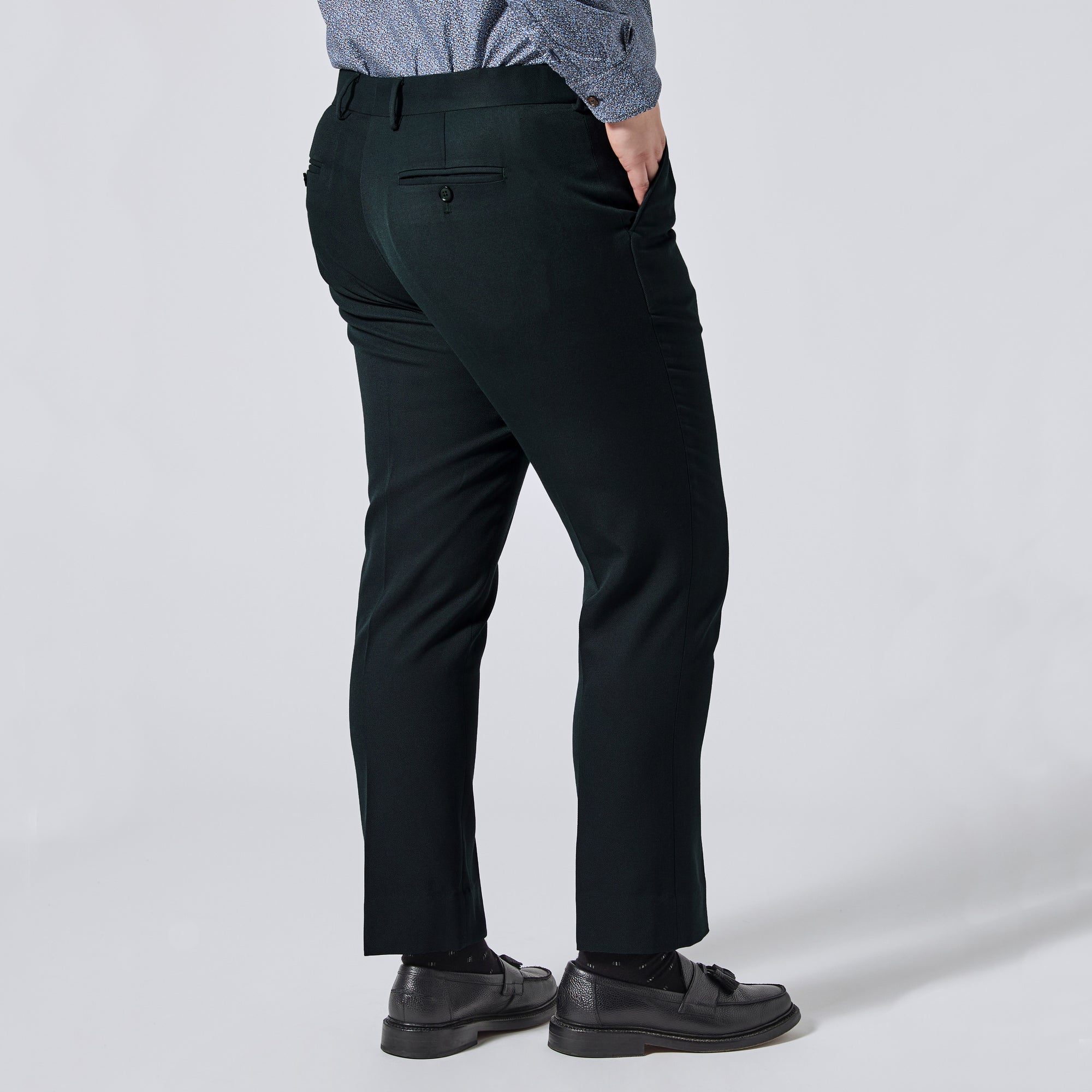 A person shown from the back wearing dark green trousers, a blue patterned shirt, and black loafers against a light gray studio background. Color: Phantom Green Twill