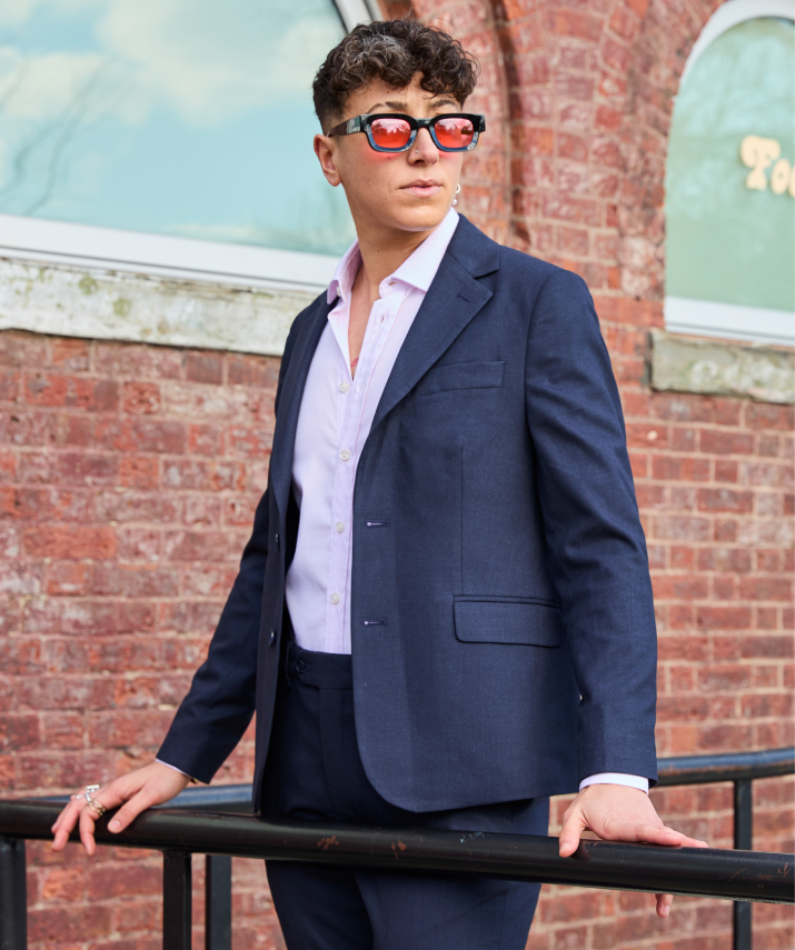 Person wearing a navy blazer and sunglasses standing against a brick wall.