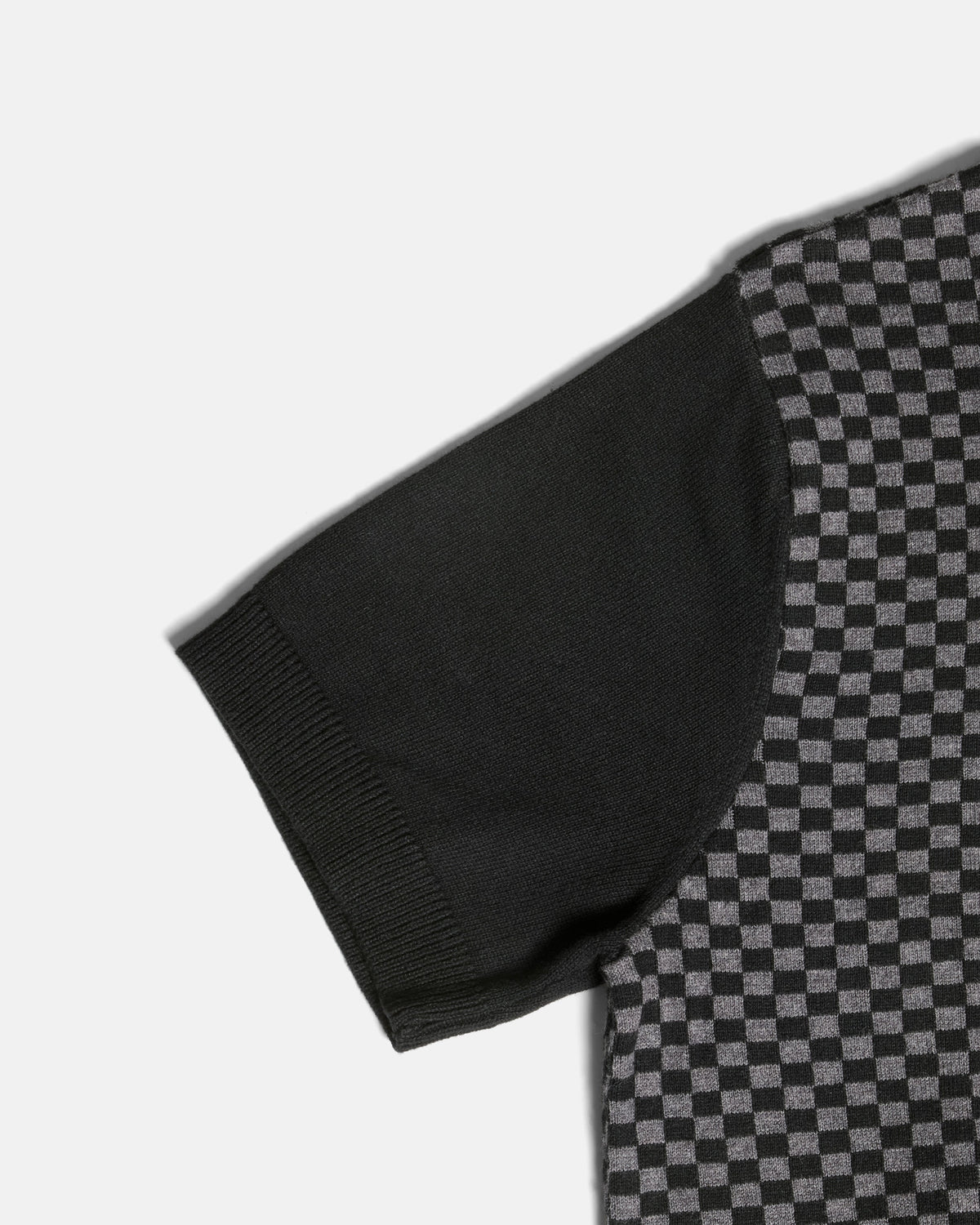 Close-up of a short-sleeve knit top showing a solid black sleeve and a black-and-gray checkerboard patterned body, photographed on a light gray background. Color: Shadow Checker