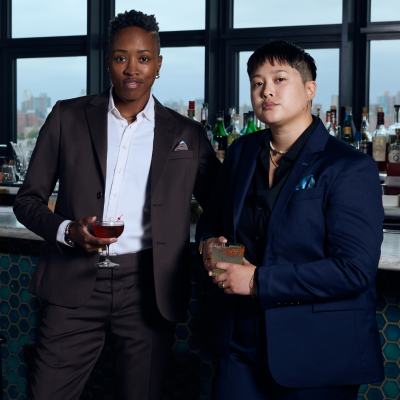 Two models wearing suits and holding drinks in a bar setting with a cityscape view.