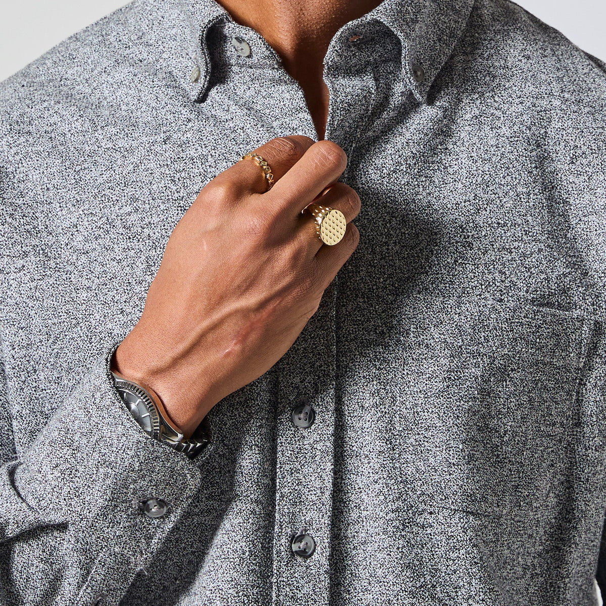 Close-up of a person wearing a textured gray long-sleeve button-down shirt, with one hand adjusting the collar and wearing gold rings and a watch. Color: Static Flannel