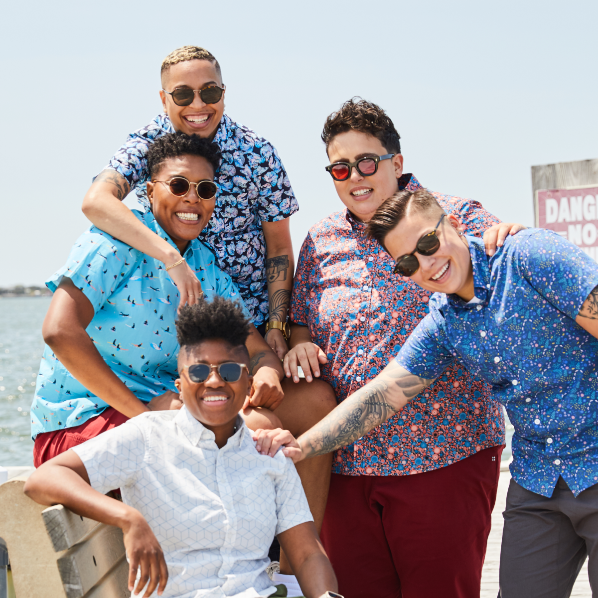 Group of five people posing together on a beach with sunglasses and short sleeve button up shirts.