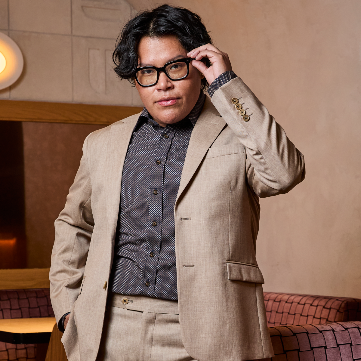 Model in a beige suit adjusting their glasses in an indoor setting
