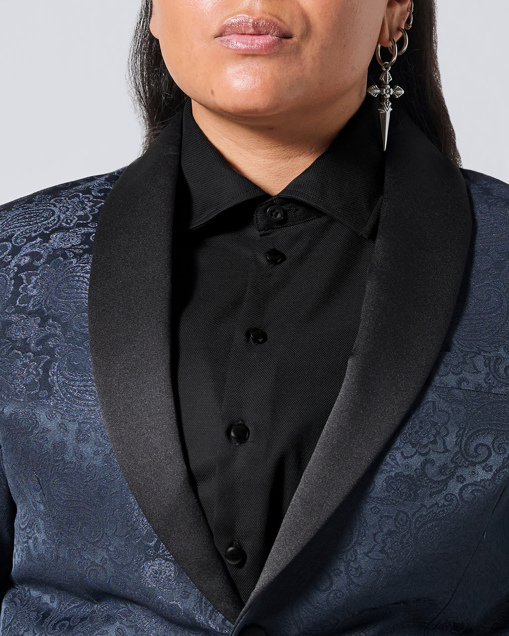 Close-up of a person wearing a dark blue patterned tuxedo jacket with black satin shawl collar over a black button-front shirt, against a light gray studio background. Color: Black