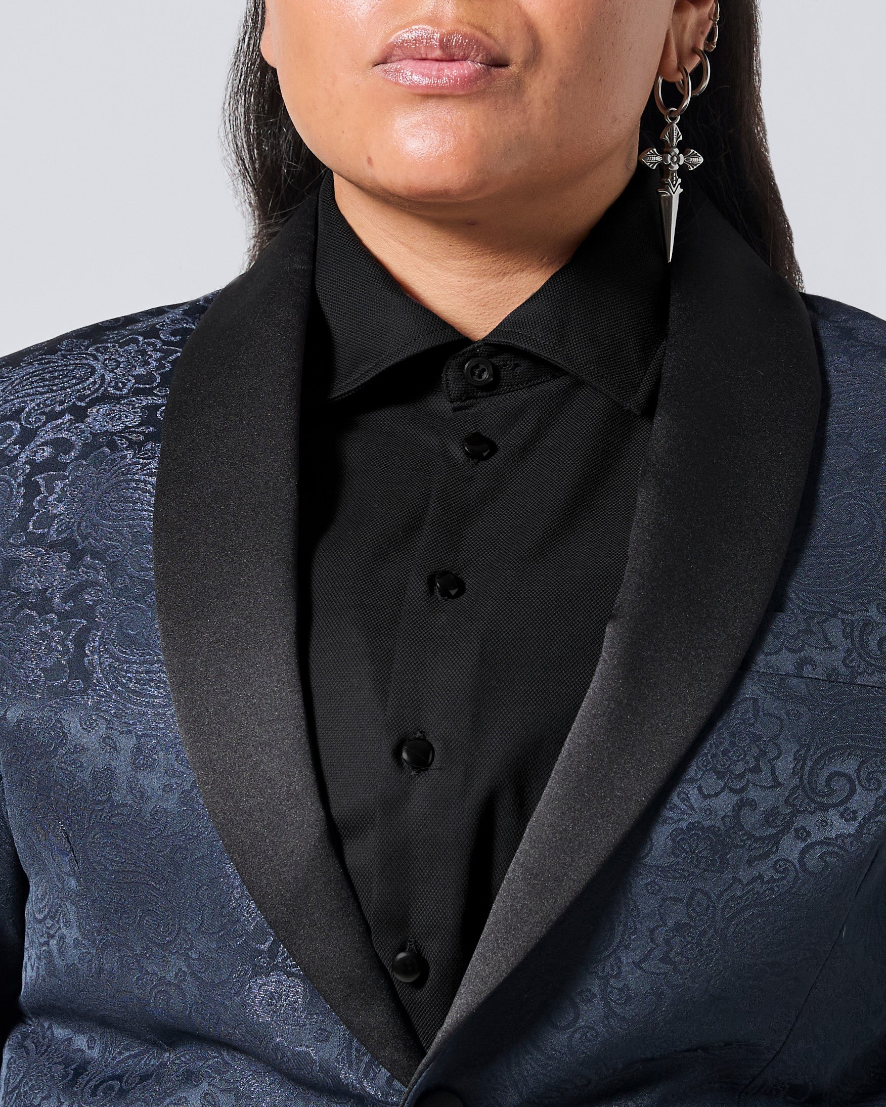 Close-up of a person wearing a dark blue patterned tuxedo jacket with black satin shawl collar over a black button-front shirt, against a light gray studio background. Color: Black