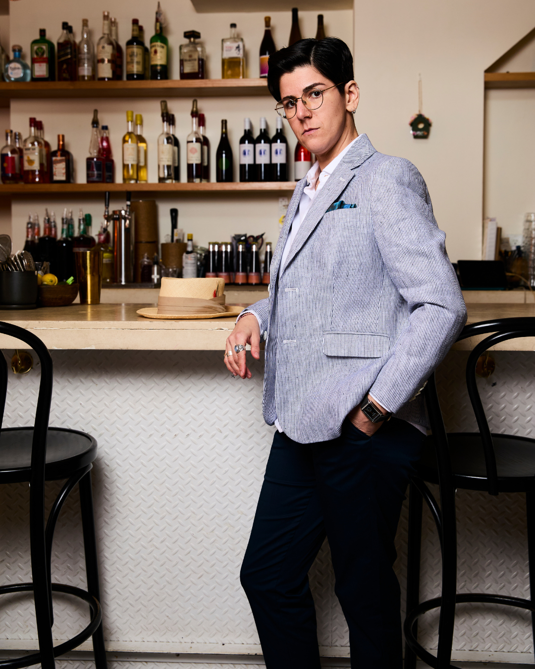 Model in a light gray blazer standing in a bar setting with shelves of bottles in the background.