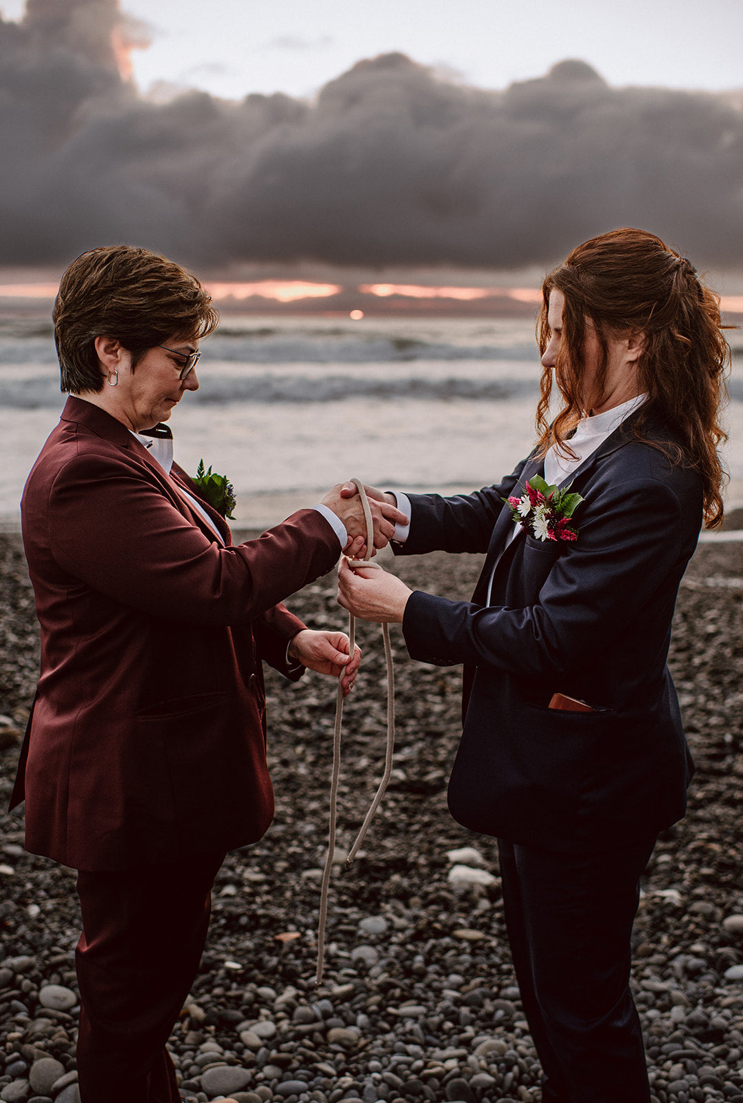 Newlywed couple in suits on a beach wrapping rope around both of their hands.