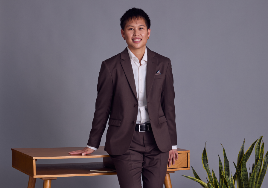 Model in a brown suit standing next to a wooden table against a gray background.