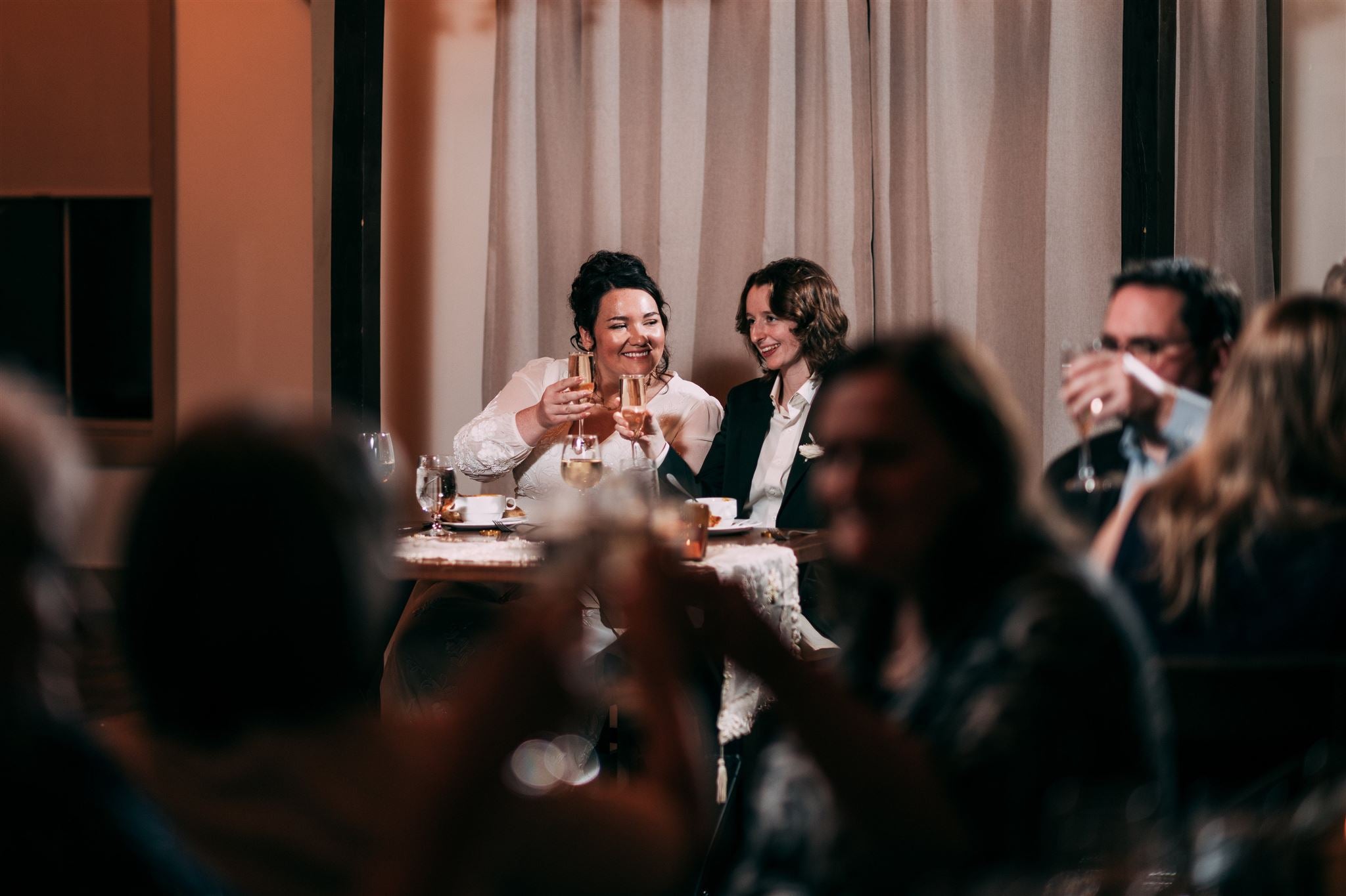 Two people sitting at a table, clinking glasses in a celebratory manner, with a blurred crowd in the foreground.