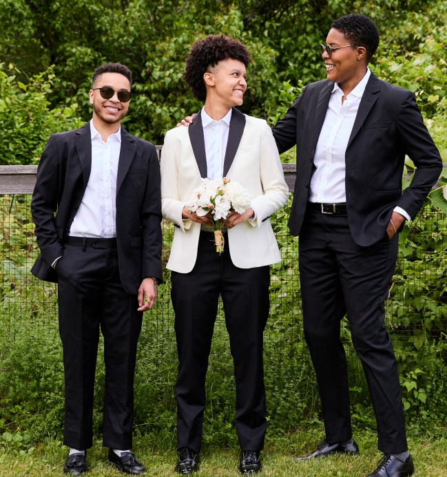 Three people in formal attire standing outdoors with greenery in the background